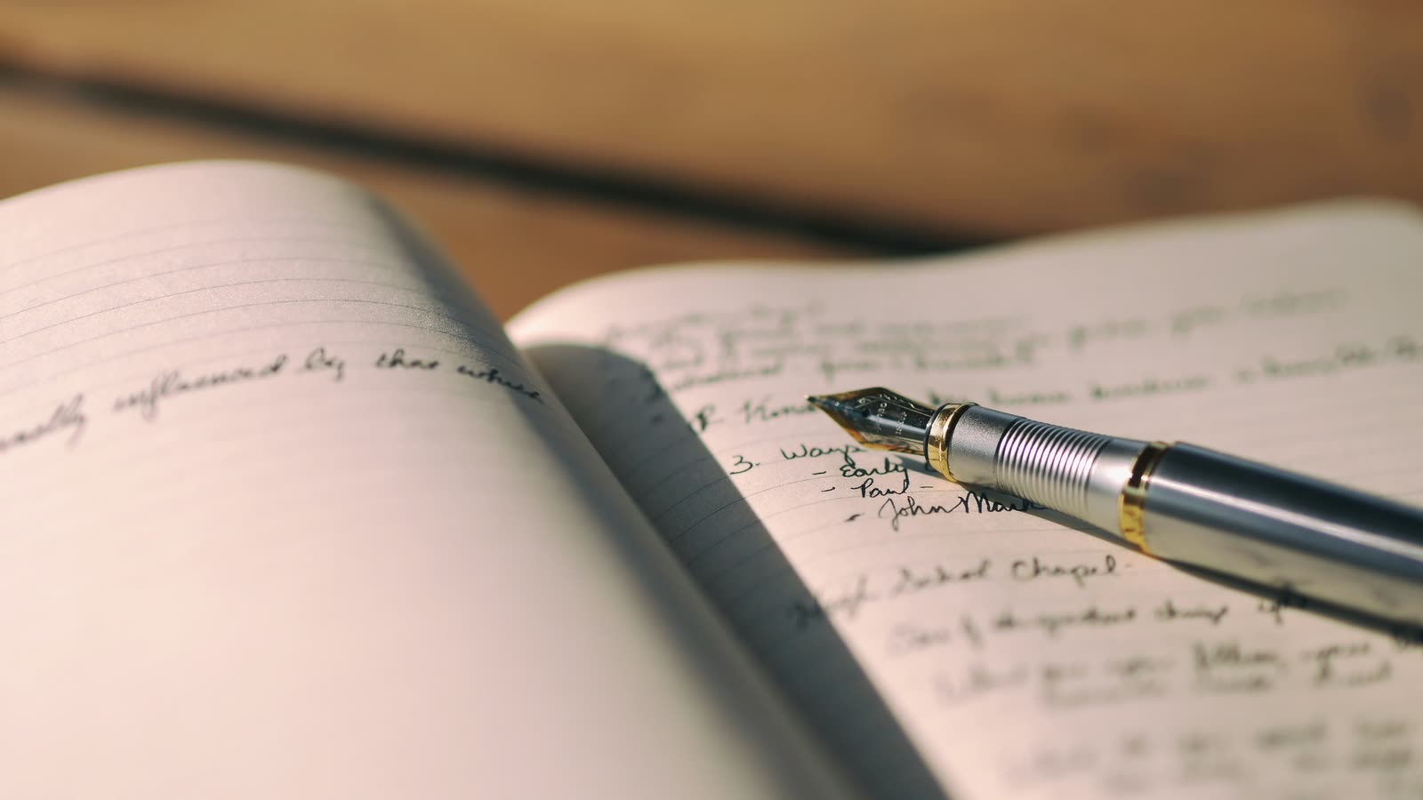 An open notebook covered in handwritten notes, with a fountain pen resting across the right-hand page on a wooden desk